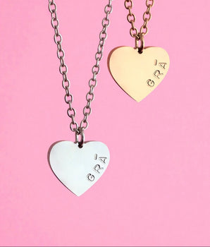 Two heart-shaped pendants on chains with 'GRA' engraving against a pink background