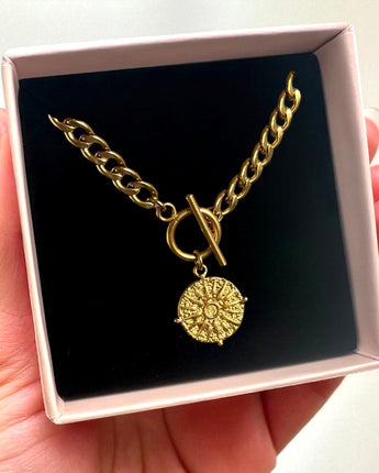 Gold chain necklace with a round pendant in a pink box held by a hand.
