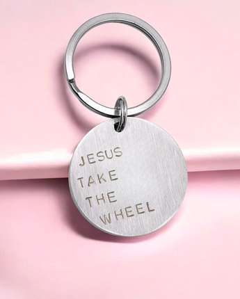 A silver keyring with the inscription 'JESUS TAKE THE WHEEL' stamped on a circular tag, attached to a pink ribbon.